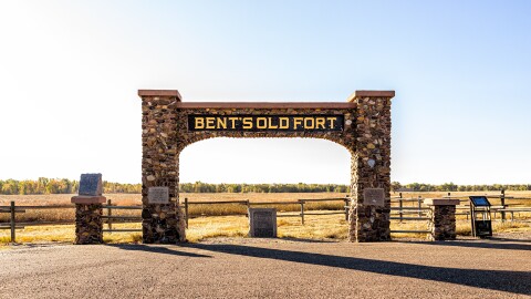 This is an image of a gated entrance with a large sign that says "Bent's Old Fort" on a clear-sky day. 