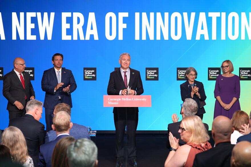 Three men and two women dressed formally stand on a stage with a sign behind them. 