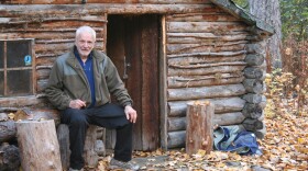 A man with short white hair sits on a log stool in front of a log cabin.