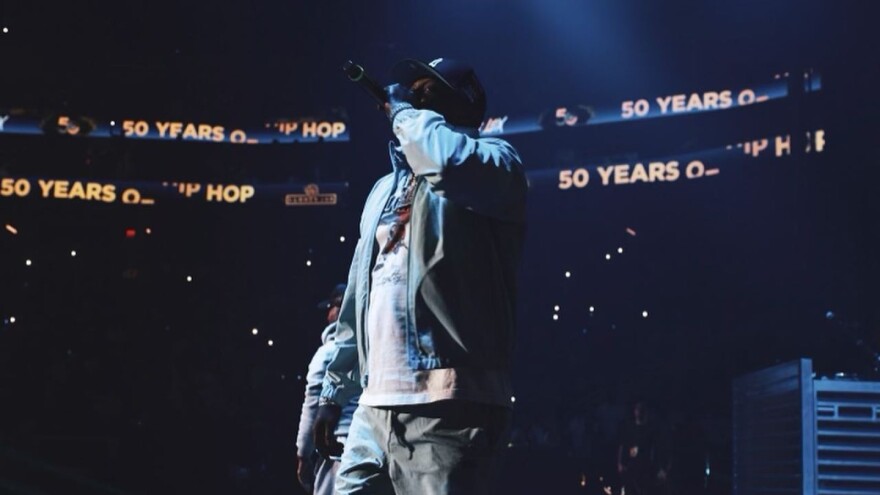 A man in a baseball cap holds a microphone to his mouth while performing on stage in a large arena with an LED ribbon board behind him that reads "50 years of hip-hop."