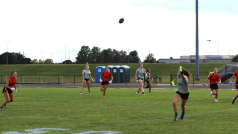 High school girls playing flag football on a grass field at a school. Several players are wearing orange and black jerseys. The others are wearing gray and green jerseys. 