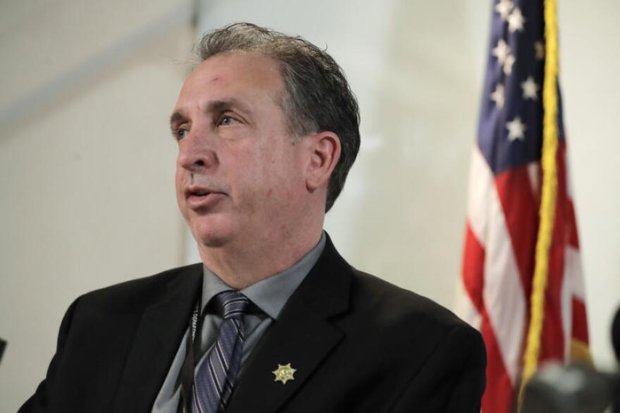 A many in a suit with a sheriff department pin speaks in front of an American flag.