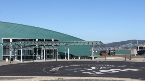 An airport terminal building overlooks a bay with a cantilever bridge in the background and a painted parking lot in the foreground with "Fly OTH" pained on it.