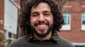 A smiling man with curly dark hair and a beard stands outdoors in front of a brick building. He is wearing a dark sweater over a green shirt with a small white pattern, and the background is softly blurred.