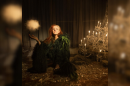 A red haired woman in a green gown surrounded by broken glass on the ground and a chandelier to the right.