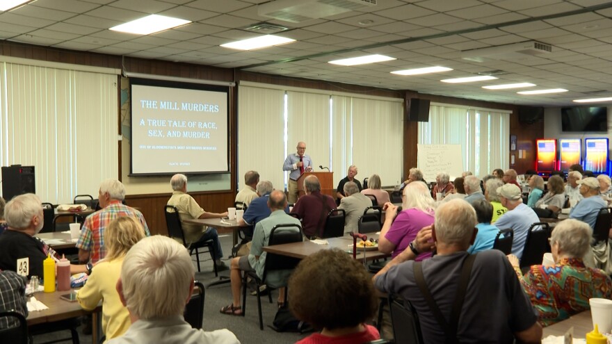 A man addresses people seated at rows of tables in front of a projector screen