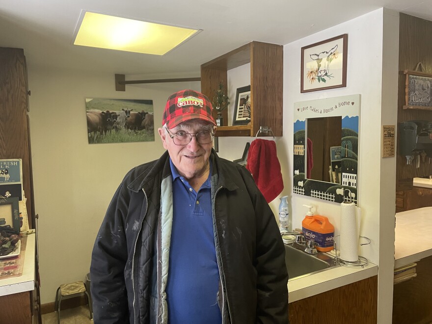 An older man in a red Cabot baseball cap and jacket stands in his mudroom. A printed photo of cows is in the background.