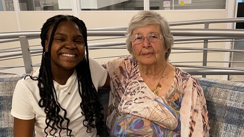 A Black teenage girl with long hair and a gray haired white woman sit together and smile