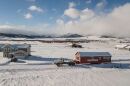 A landscape photo of a snow covered valley. A yellow two story house with a garage is n the left of the frame. And a tow truck pulling a long red house is in the right part of the frame. 