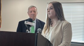 Dentist Anne Koehne speaks at a news conference with U.S. Senator Dick Durbin standing in the background. There is a podium with a WGLT microphone on it.