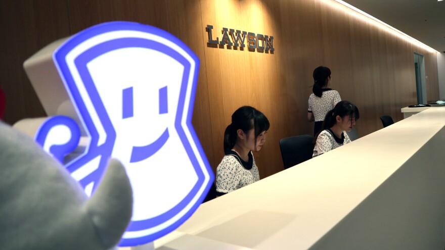 The front lobby of Lawson company headquarters in Tokyo. A cartoon version of the Lawson emblem sits on a long white counter. Three women wearing matching black and white dresses sit behind the counter.