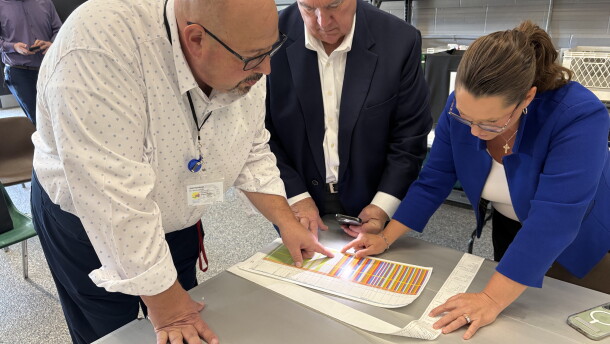 Three people lean over a table, looking closely at a sheet of paper with colored lines.