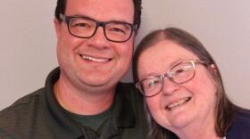 David and Cindy  Cordero side hug at their StoryCorps interview. David stands at least half a head taller than his mother, who leans her head in toward his shoulder. David has short dark hair and wears dark framed glasses and a dark heathered gray pullover.  The pullover is slightly unzipped and he is wearing a black tee shirt underneath it.  Cindy has light brown strawberry hued hair that falls to her shoulders.  Wisps of gray fall in front of her ears.  She has cat's-eye glasses.  She wears a blue printed white tee shirt and a dark cardigan.