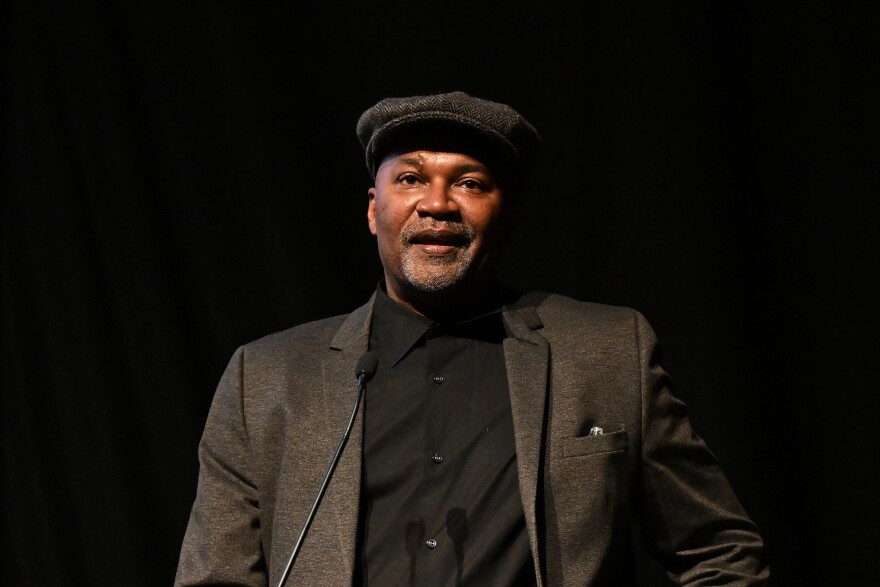 A man wearing a brown suit and cap speaks into a microphone at a lectern while standing in front of a dark background.