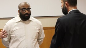 A man wearing handcuffs stands before a man in a suit in a courtroom.