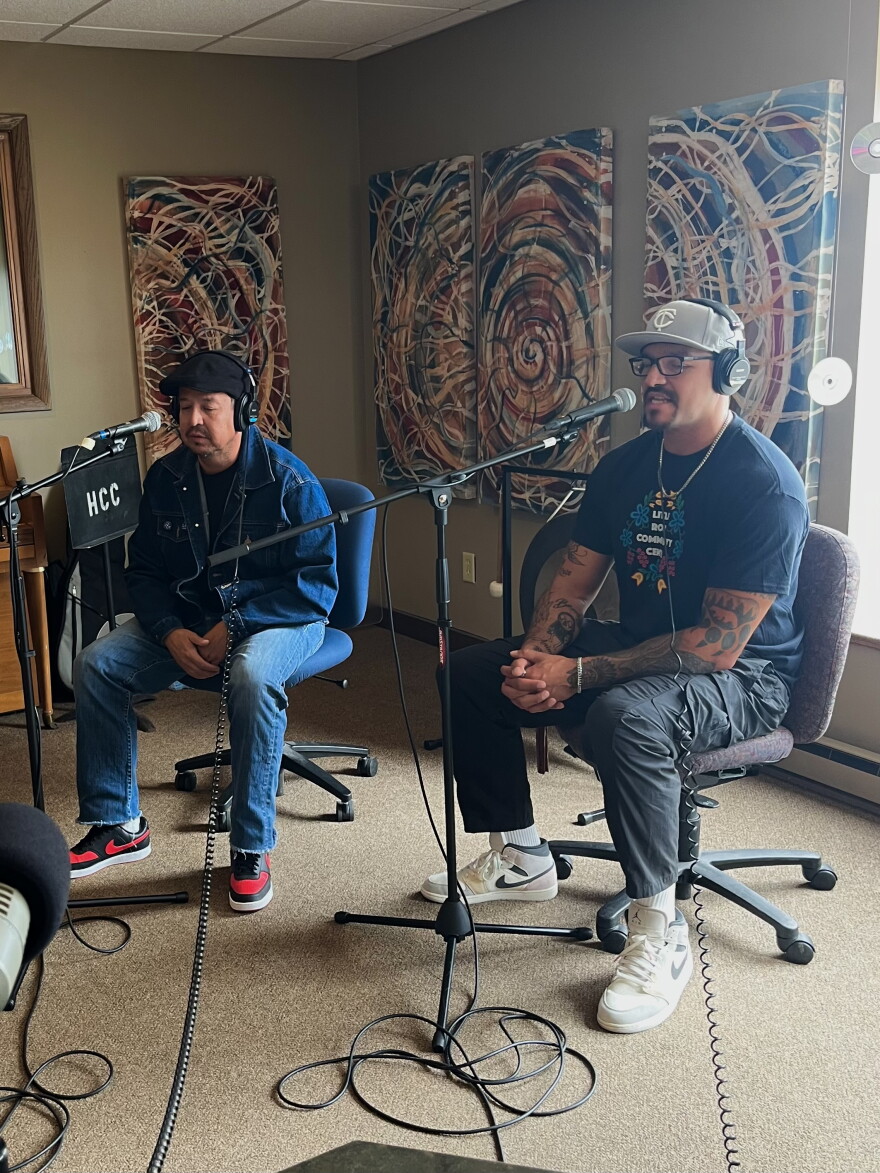 Two men sitting behind microphones in a radio studio. One is rapping into his microphone.
