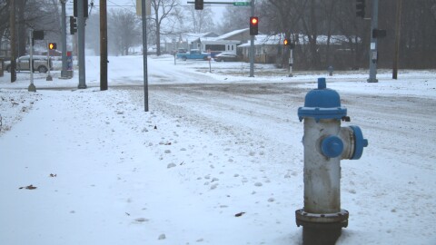 A blue and silver fire hydrant is in the right side of the frame, snow surrounds it. In the background are traffic lights and an intersection. The roads are snow covered.