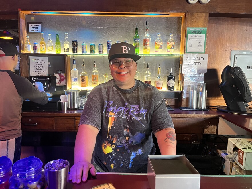 Patty stands behind the bar at the White Horse wearing an Oakland Ballers cap and a Prince t-shirt, smiling.