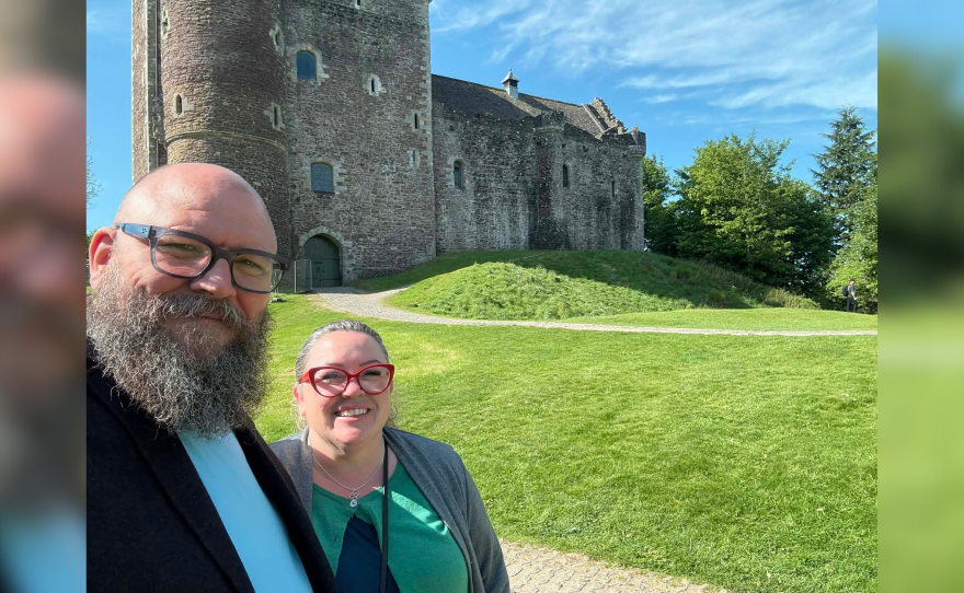 Two people posing for a picture in front of grey castle with a lot of green grass surrounding it.