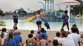 Three adults stand in front of a small crowd of children and lifeguards in pool clothes. The adults on either side adjust their life jacket fit, while the adult in the middle faces the crowd to engage with the kids, her arms outstretched as she asks a question. Behind the presenters is Juniper Hill pool, with a children's pool play area in the immediate background.