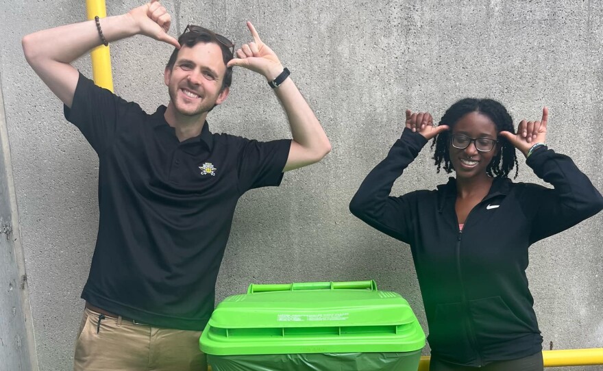 A man wearing a black polo shirt and khakis and a woman wearing black leggings and a black hoodie, stand by a waist-high neon green compost bin