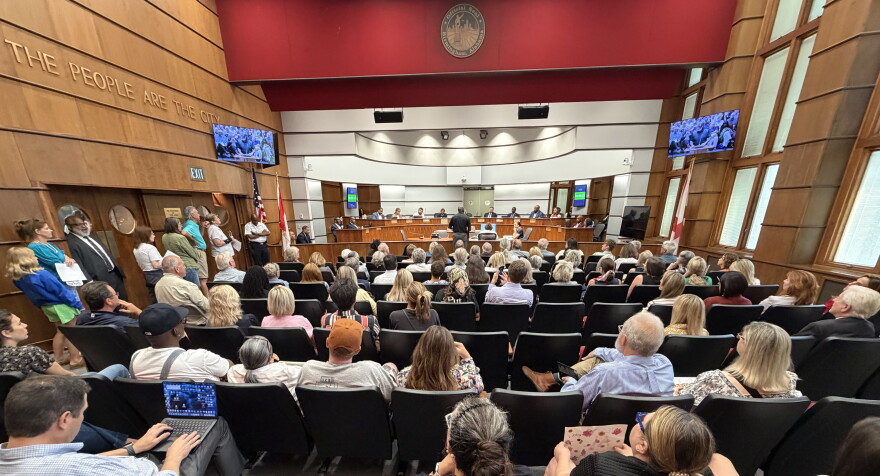 People fill seats and stand in line along one wall at a meeting in the Birmingham City Council's chambers.