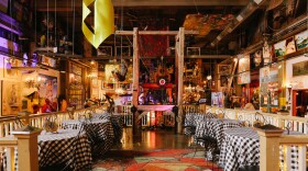 A room filled with various art and objects, including paintings, tables with black and white checked cloths, chairs, a colorful rug, and different lights