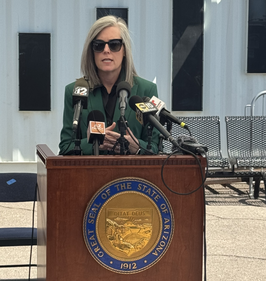 Woman in sunglasses and green jacket speaks at wooden podium