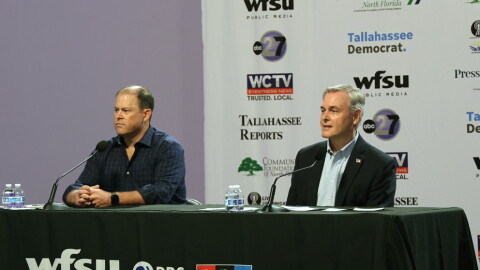 Two men sit at a table in front of microphones staring forward.