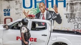A police officer stands by a wall painted with a mural of late Haitian President Jovenel Moise. (Photo by Valerie Baeriswyl / AFP)