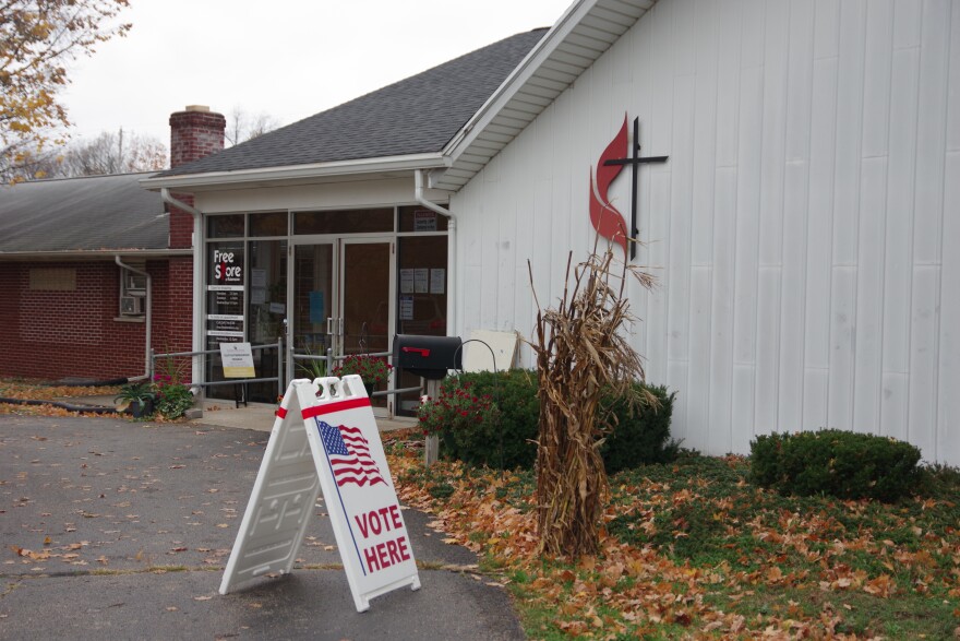 A small white sign that says Vote underneath an American flag. A stalk of corn stands next to it. Behind it is a church that is half brick and half white. There is a black cross with a red flame on the church. 