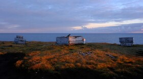 Ice cellars, constructed of wood boards, rest on a grassy field with the ocean looming in the background.