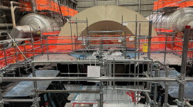 Metal scaffolding and bright orange netting cover the room, while the light-brown metal arch of the partially dissassembled turbine cover sits inside the Palisades nuclear plant.