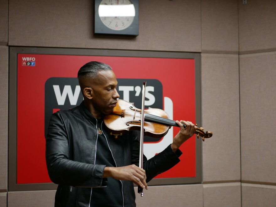 A man in a black jacket plays the violin in front of a red sign that says "What's Next?"