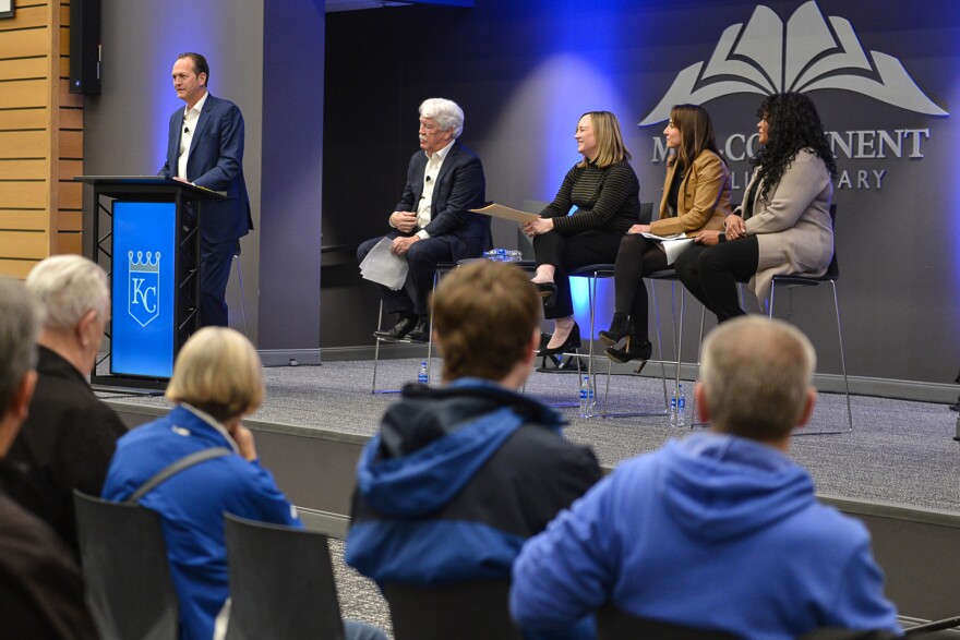 A man wearing a suit talks at a podium on a stage in front of a sign that reads "Midcontinent Library." Four other people are seated to his right. Audience members sitting in chairs in front of him.