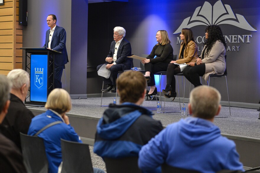 A man wearing a suit talks at a podium on a stage in front of a sign that reads "Midcontinent Library." Four other people are seated to his right. Audience members sitting in chairs in front of him. 