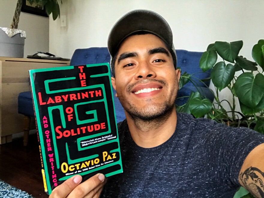 Miguel Perez sits in front of a plant in his home holding the book 'The Labyrinth of Solitude'. 