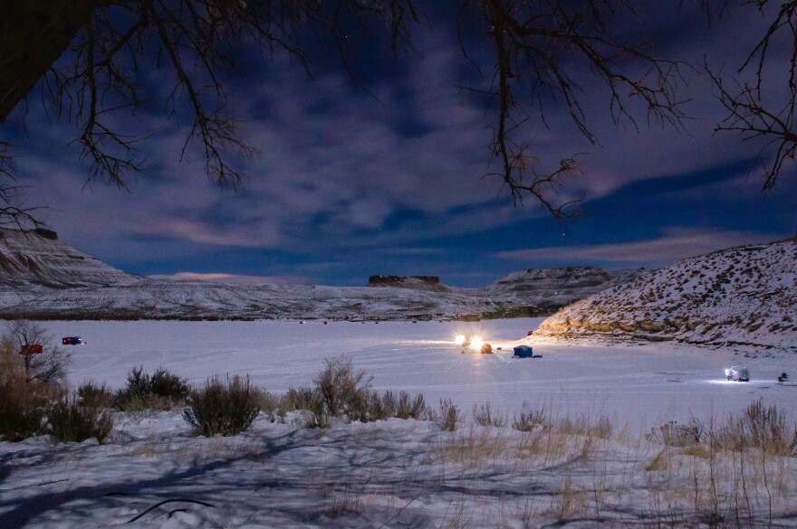Tents and lights are scattered around a frozen lake, with tree branches in the foreground and snowy hills in the background on the far edge of the lake.