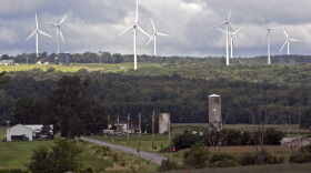 A rural landscape. Farms and trees are visible in the foreground. In the background are 8 large white windmills.