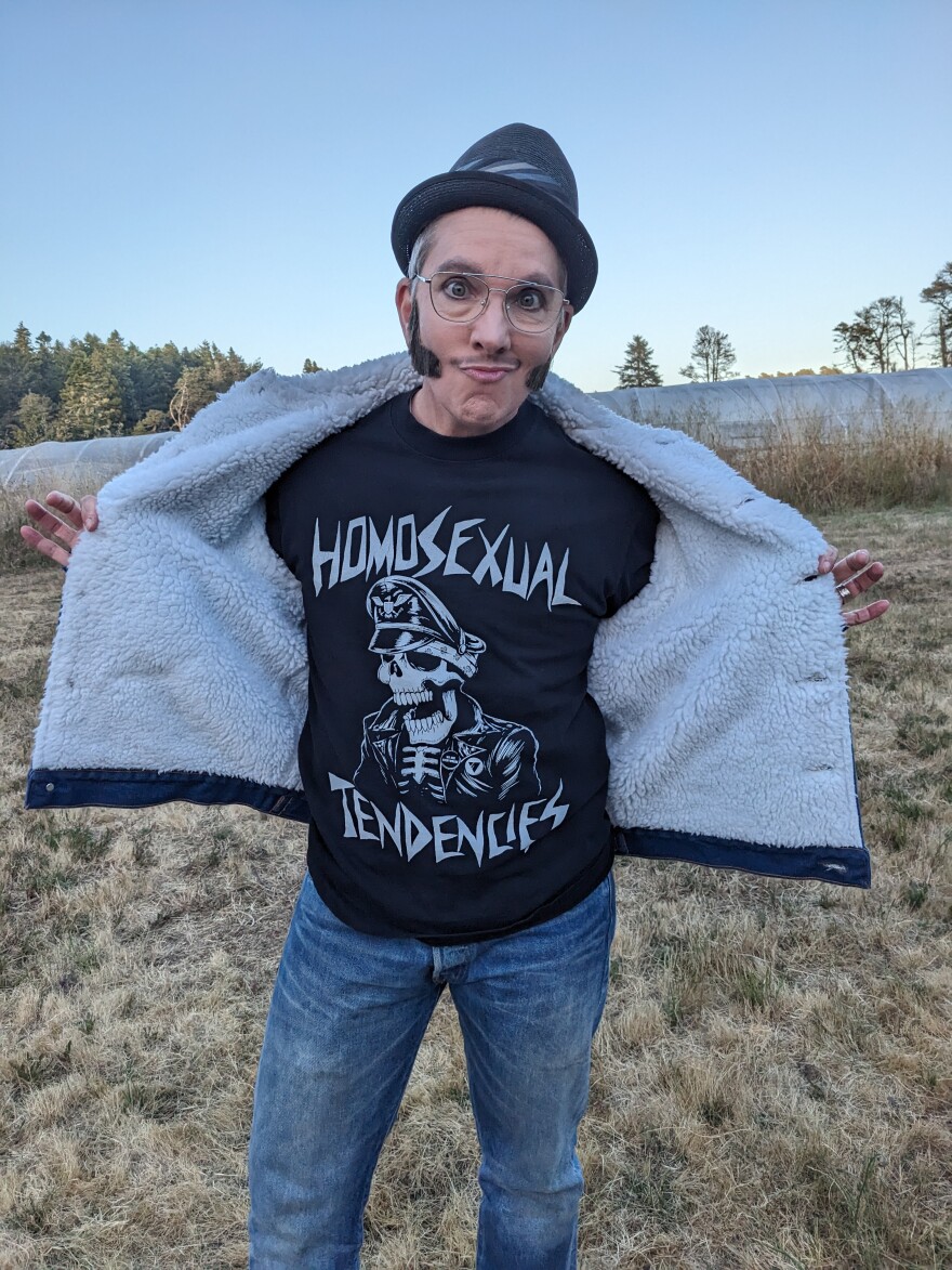 A performer in fake sideburns holds open a fleece jacket to reveal a T-shirt that says "Homosexual tendencies."