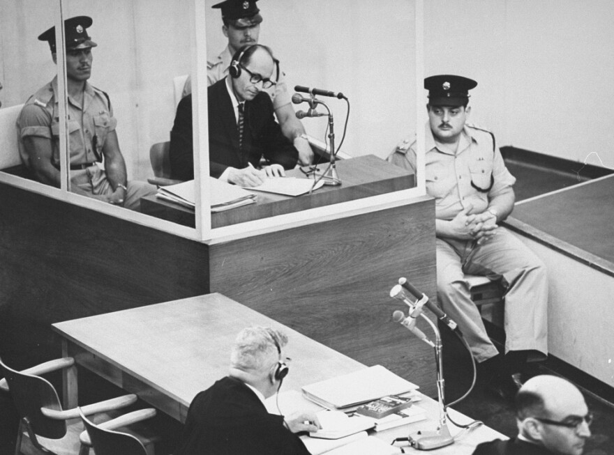 Defendant Adolf Eichmann takes  notes during his trial in Jerusalem.  The glass booth in which Eichmann sat was  erected to protect him from assassination.