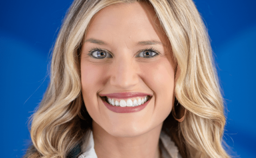A smiling woman with long blonde hair is wearing a green jacket over a button-down blouse.