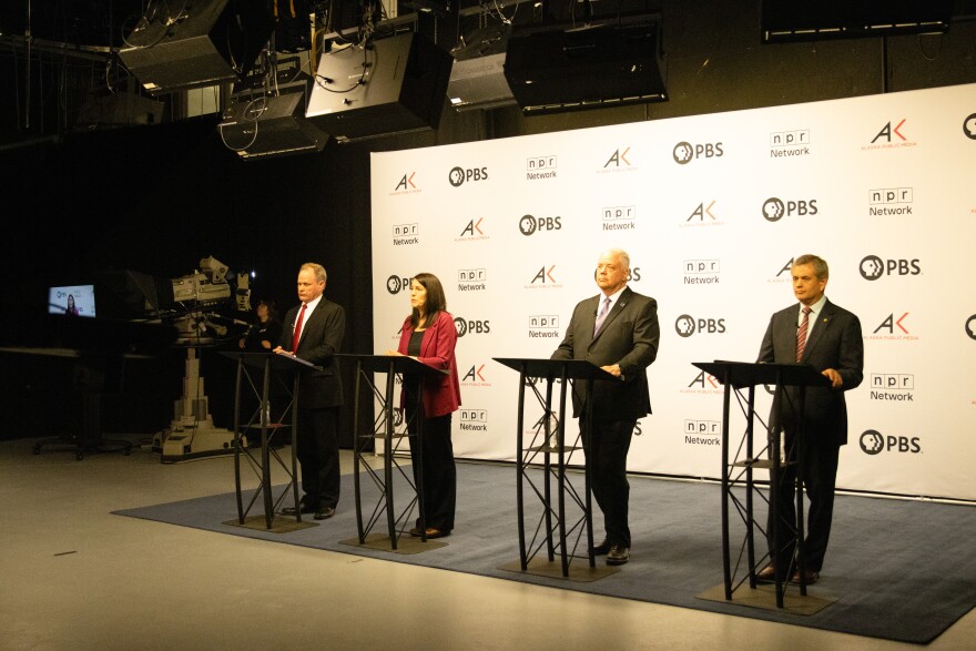 Four people, a man in a suit with red tie, a woman with a deep red blazer, a man in a suit with purple tie, and a man in a suit with a striped tie stand behind podiums in front of a white backdrop with the logos for PBS, NPR, and Alaska Public Media preparing for a debate.