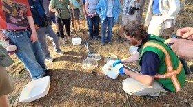 people of various ages stand in a semi-circle around a man in green vest with a tub of water and salamanders