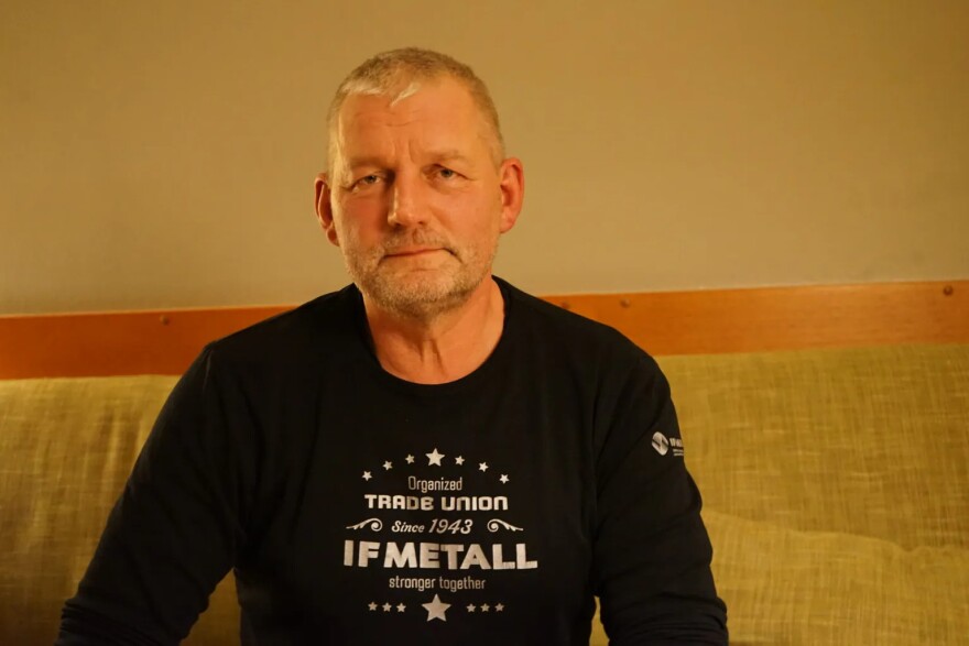 A white man sits on a couch while wearing a black long sleeve shirt with "Organized trade union since 1943 IF METALL stronger together" written on it.