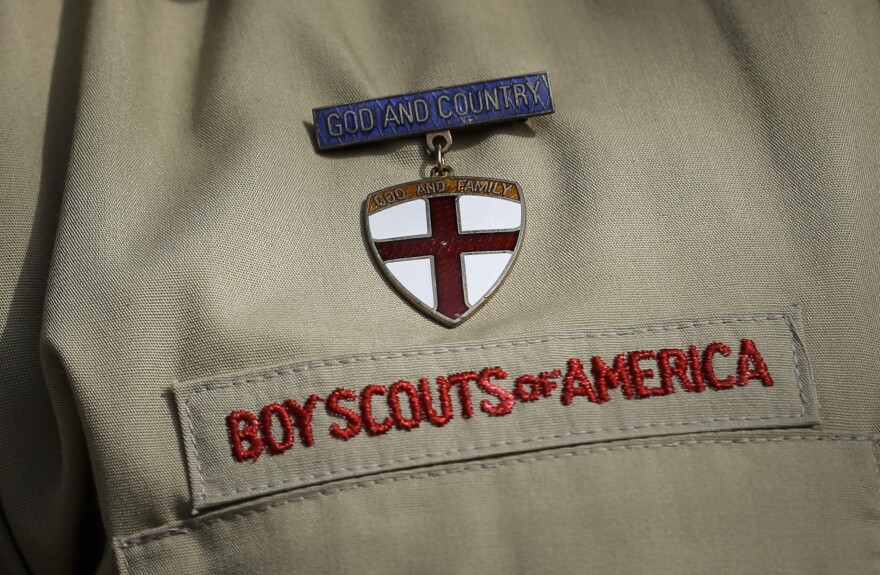 a shield-shaped badge with red cross on white background hangs from a blue pin reading "god and country" on a tan uniform, embroidered with the words "Boy Scouts of America" in red