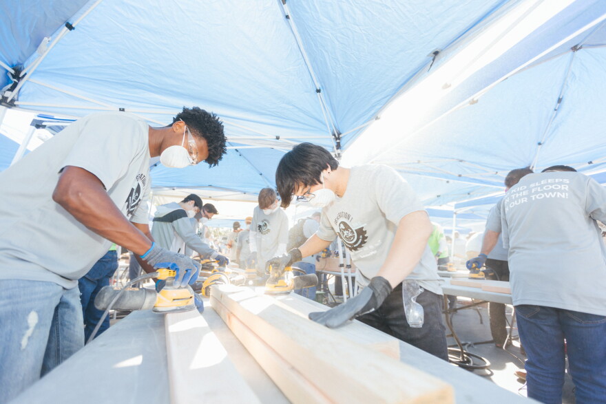 Volunteers wearing masks and safety glasses use power sanders on lumber beneath outdoor canopies.