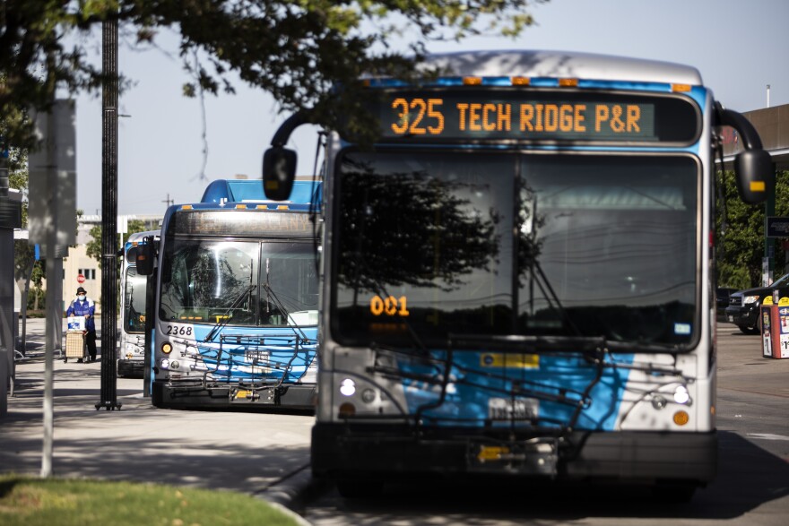 Capital Metro buses lined up at Norwood Transit Center. Each bus is parked at the curb waiting for passengers. In the distance, a man wearing a blue mask and a black fedora is pushing a personal cart containing some belongings along the sidewalk towards the buses. 