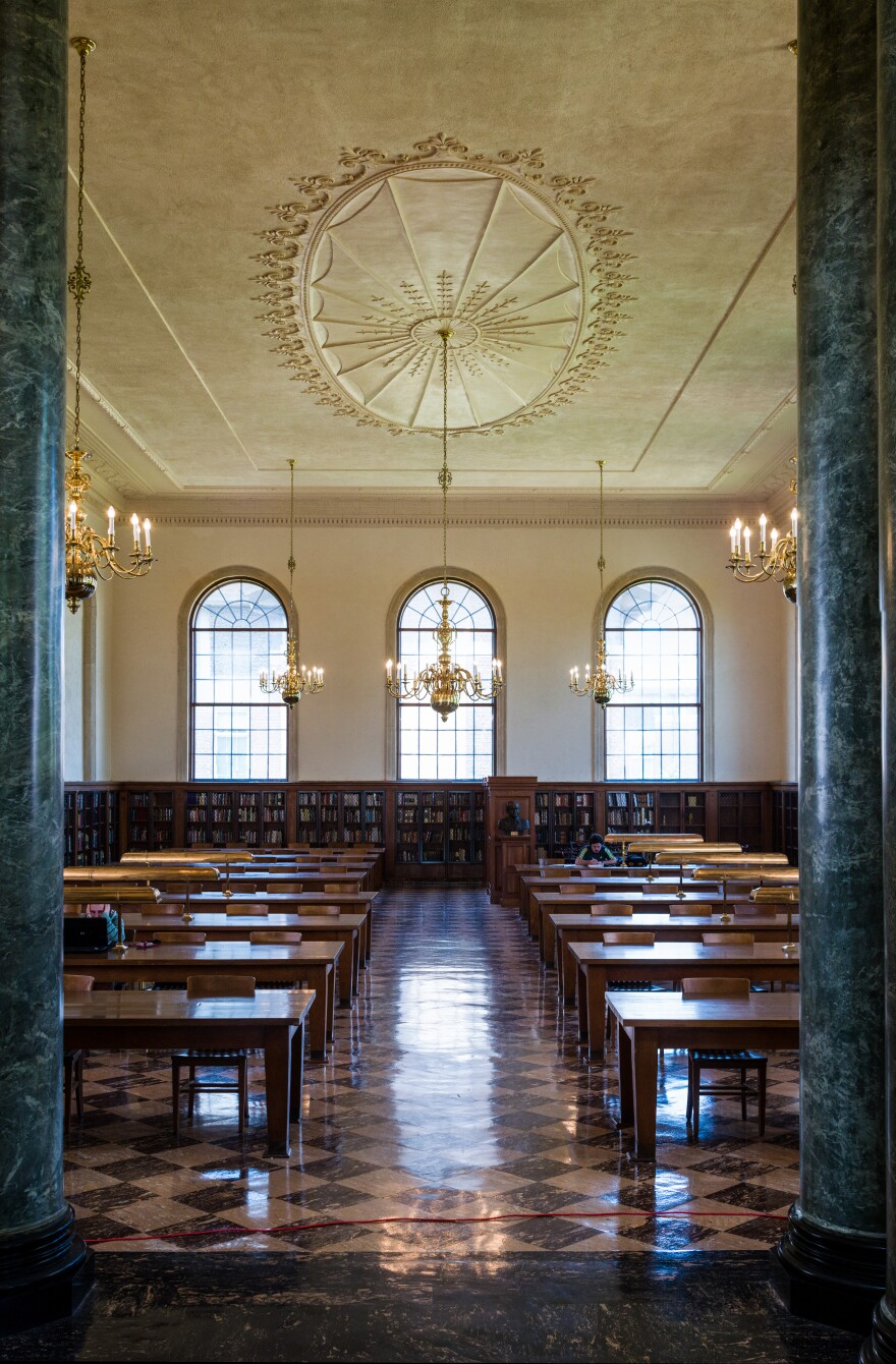 A view of the main reading room in Wilson Library. Wooden tables sit in rows on a black and white checkered floor. Green columns act as a gateway into the room, lit by arched windows and chandeliers hanging from a decorative ceiling. 