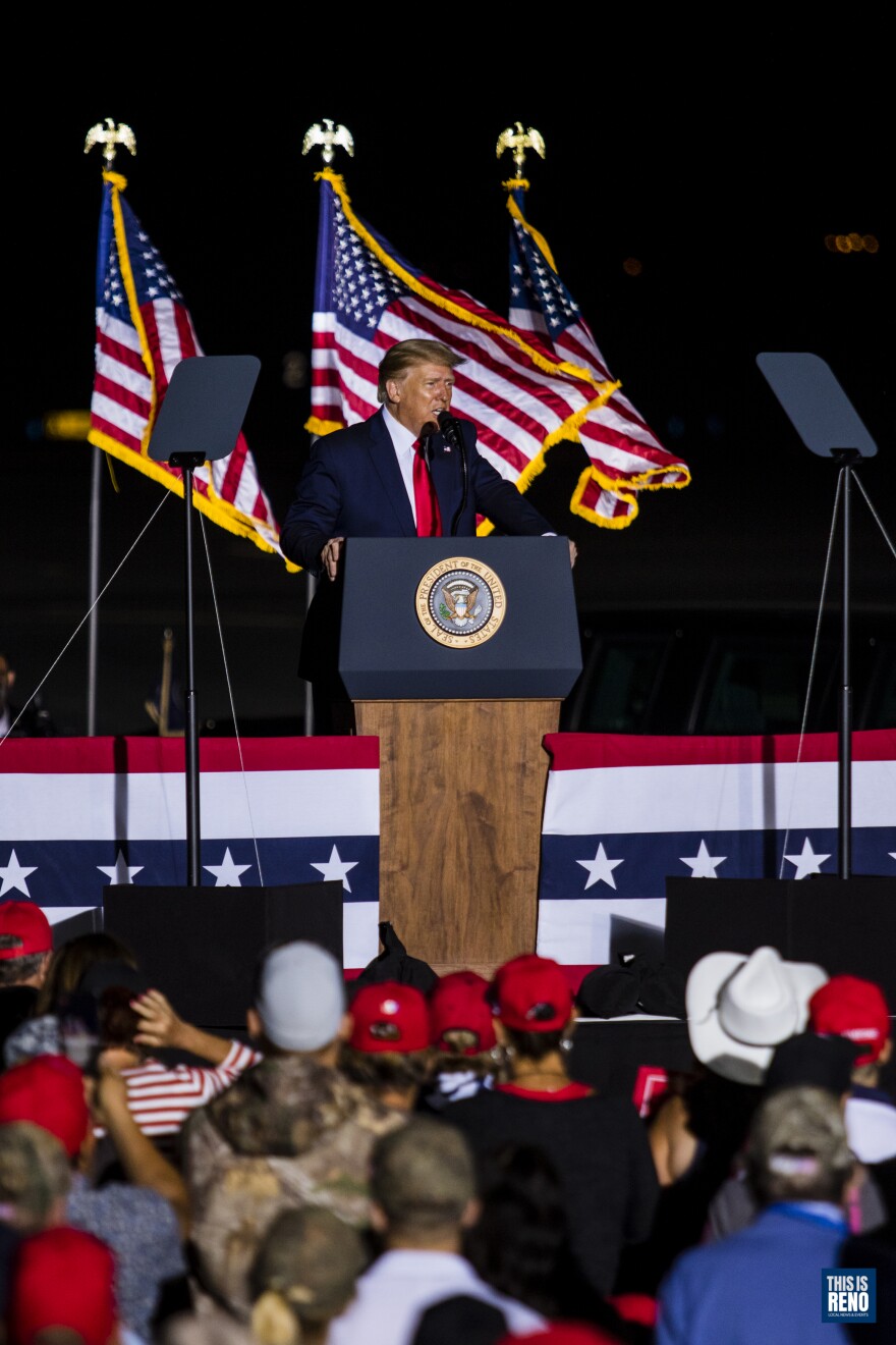 Its dark outside. A vertical photo of a man behind podium with three American flags behind him. He’s in front of a crowd.